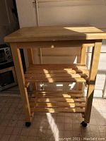 Front view of wooden butcher block kitchen cart with two open slatted shelves and caster wheels on a tiled floor.