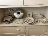 Wide shot of various plates and platters arranged on a counter including a metal lid and ceramic pieces with floral designs.