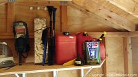 Wide view of upper shed shelf showing red plastic fuel cans grouped with window scrapers, sun shields, small black funnel, and VP small engine fuel can.