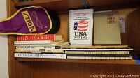 Wide shot showing the full lot of books stacked on a wooden shelf alongside one Stiga ping pong paddle in cover.