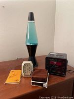 Photo showing lot on wooden surface including blue liquid lava lamp, octagonal Citizen clock, Soundesign black digital clock radio, suede pouch, and metal note pad holder.