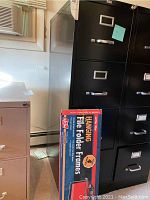 Black Staples 4-drawer file cabinet with some scratches and a box containing two hanging file folder frames in the background.