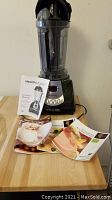 Photo showing the Health Master blender with clear jar, lid, black base with control buttons, owner's manual, and recipe book on a wooden surface.