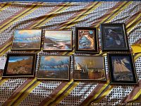 Photo showing all eight framed holographic pictures arranged on floor with striped textile background. Includes beach, boat, eagle, clock, cottages, sailboat images. Frames show glue splitting and wear.