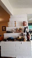 Overall display of the lot items on a white brick ledge, showing various model cars, candlesticks, ceramic pot, wooden wagon, bronze figurine, and decanters.