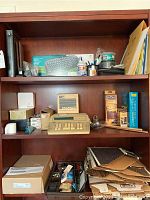 Wide view of wooden shelving unit holding multiple office items including vintage adding machine, binoculars, keyboard, stationery, and books.
