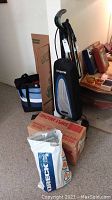 Oreck upright vacuum cleaner with hose and bag standing upright beside a box of Oreck vacuum bags and an Oreck canister vacuum in its box on floor