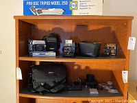 Overview of Polaroid OneStep Plus camera, Nikon D100 camera, Nikon camera bag, Minolta camera box and other items arranged on a wooden shelf.
