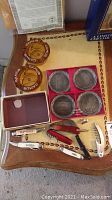 Photo showing 2 brown Canada Games 1977 ashtrays, 4 metal Baseball Hall of Fame coasters in red velvet case, various pocket knives and cigar accessories on a wooden table.