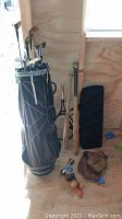 Photo showing black and gray golf bag standing upright with multiple golf clubs, two baseball bats, two worn gloves, horseshoes with balls, racquet balls, weights, and massage tool on the floor in a wooden shed.