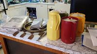 Three vintage pitchers in red, yellow, and orange colors on countertop with metal serving tray and linens