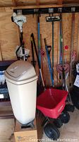 White plastic bin and red broadcast seeder in front of assorted long-handled tools, Stihl saw mounted above