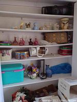 Closet shelves with Honeywell heater on top, various ceramic and glass figurines, pink and clear glass decanters, decorative tins, plastic storage containers, artificial flowers and miscellaneous items in boxes.