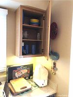 Wooden kitchen cabinet open revealing shelves with colorful bowls and drinking glasses, box of Tempreseve item and two cutting boards on countertop along with a toaster covered in a white cloth.
