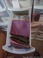 Open plastic bin drawer showing folded quilting fabric pieces in various colors and patterns including purple with small dots, brown, reds, blues.