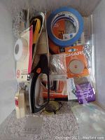 Various quilting tools inside a box including rotary cutter, masking tape, Stitch Witchery, Skars rotary cutter blade, blue painter's tape, and quilting ruler.