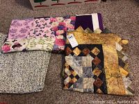 Photo of three folded quilts on carpet, showing different colorful patchwork patterns including beige, brown, purple, butterfly prints, and florals.
