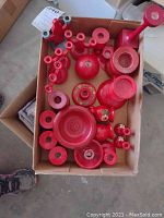 Overview of all red wooden candleholders and decorative pieces arranged inside a cardboard box showing variety of shapes and sizes.