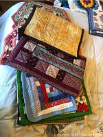 Stack of several handmade quilted table runners showcasing a variety of patterns including patchwork, floral, and geometric designs in multiple colors