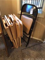 Six wooden TV trays and one metal folding chair leaning against a wall on carpeted floor near two windows.
