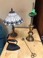 Shows Tiffany-style stained glass lamp, brass finish lamp with no shade, vintage cast iron flat iron, and metal candle snuffer on wooden table.