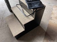 Three-step dog stairs with beige carpeted steps, dark brown wooden frame, shown from an angle with the black leather purse on top.