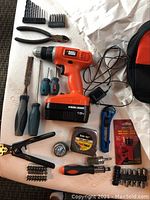 Full view of Black and Decker cordless drill, pliers, screwdrivers, measuring tape, utility knife, wire brushes, bit racks, and charger cord on table.