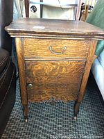 Front view of oak side table showing drawer with metal handle and cupboard door below with metal knob. Clear view of wood grain and carved details on legs and bottom edge.