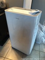 Front view of white Danby portable air conditioner with remote control resting on top, unit standing on tile floor.