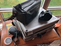 Camera and accessories neatly placed on and around aluminum carrying case on wooden table by window with natural light. Includes black leather bag, various filters, cables, and small cases.