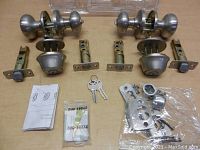 Two sets of Kwikset locking door knob sets each with door knobs, deadbolts, keys, strike plates, screws, and manuals on a beige fabric surface.