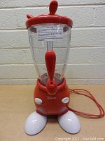 Front view of red and white Disney Mickey Mouse themed smoothie maker with clear jar, red lid with Mickey ears, dispenser spout and Disney logo on base.