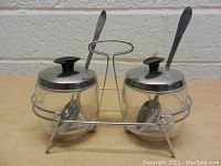 Front side view of the two glass containers with stainless steel lids and spoons in the wire rack against a brick wall background.