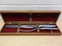 Top-down view of carving knife and fork inside open walnut wooden case lined with red fabric.
