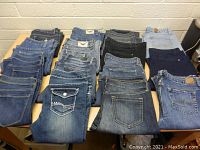 Wide view of 15 pairs of women's jeans folded and arranged on a table showing various washes and colors from light to dark blue.