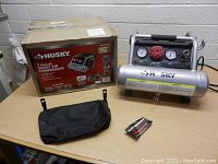 Photo showing the Husky 1 gallon silent air compressor, original box, and black carrying/storage bag on a wood surface.