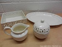 Front angled view of cream pitcher, sugar bowl with lid, napkin holder, and oval platter showing ivory lace cut-out pattern detail.
