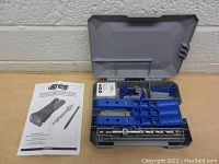 Open gray plastic case containing blue Kreg jig components, a large drill bit with adjustable collar and two rods, and additional blue plastic accessories. White Kreg Pocket Hole Jig Owner’s Manual is placed beside the case.