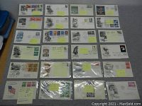 Top-down photo showing a complete view of 25 vintage first day issue envelopes arranged in rows on a flat surface, enclosed mostly in protective plastic sleeves. Various colorful stamps and cancellation marks are visible.