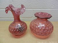 Photo showing two pink hobnail glass vases, one tall with ruffled top and one shorter with flared ruffled rim, on a wooden surface against a light wall.