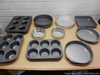 Photo showing assorted baking pans including muffin pans, circular cake pans, springform pans, a square baking pan, and an oblong baking pan on a wooden surface.