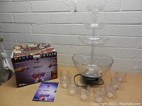 Party fountain assembled on table with punch bowl lit from base, accompanied by 10 clear plastic glasses, original box with colorful party design, and instruction manual.