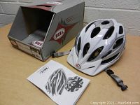White BELL adult bike helmet with multiple vents, adjustable grey straps, original box and user manual on a wooden surface.