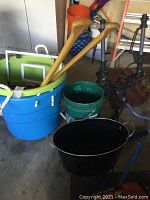 Photo showing the black metal bucket, wooden oars, three colorful plastic buckets stacked inside each other, an orange plastic bucket on top, and other assorted items in background.