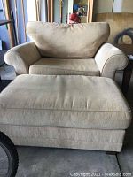 Full view of beige upholstered chair with cushions and wide armrests next to ottoman on concrete floor in garage setting.