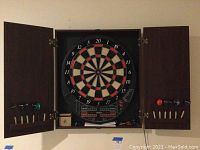 Electronic dart board housed inside a wooden cabinet with doors open showing the darts and board.