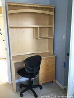 Desk and chair placed in a corner against blue walls, showing the full desk with hutch and three drawers beneath and the black office chair in front