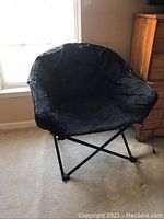 Full view of black fabric bucket-style folding chair with metal criss-cross legs on carpet flooring in front of window.