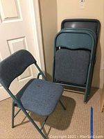 Photo of one blue fabric folding chair unfolded and four chairs folded and stacked against the wall.