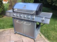 Full view of stainless steel Dyna-Glo propane grill with six burners, side burner on right, storage cabinet below, set outdoors on patio.